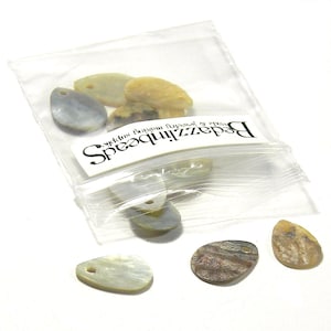 May include: A bag of white, brown, and grey shell pendants with a hole drilled through each one. The bag has a label that reads "Beadazzled" and "Spectacular Bijoux & more".