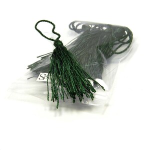 May include: A package of dark green tassels, each with a looped top and a long, fringed body. The tassels are made of a shiny, possibly synthetic material. The package is clear, and the background is white.