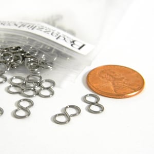 May include: A close-up of silver-toned metal jump rings, also known as split rings, in a clear plastic bag. The jump rings are about the size of a US penny, which is shown for scale.