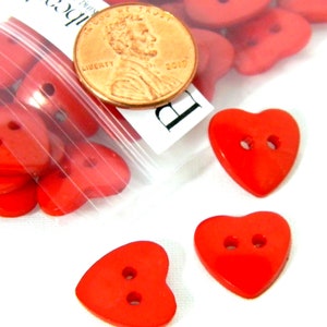 May include: Bright red heart-shaped buttons with two holes, scattered on a white surface. A clear plastic bag contains more buttons. A US penny is placed on top of a small white tag.