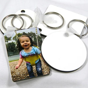 Puede incluir: Llaveros con anillas metálicas. Un llavero presenta una fotografía de un niño sonriente en un columpio. Otros llaveros están en blanco, con una superficie blanca y bordes negros. Los llaveros son rectangulares, circulares y cuadrados.
