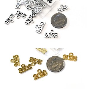 May include: Silver and gold-colored 2026 charms, some in small plastic bags, are displayed next to a dime. The charms are small, with a loop at the top for attaching to jewelry or other crafts. The background is white.