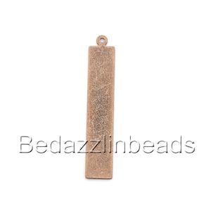 May include: A long, rectangular, antique brass charm with a textured surface and a loop at the top for hanging.