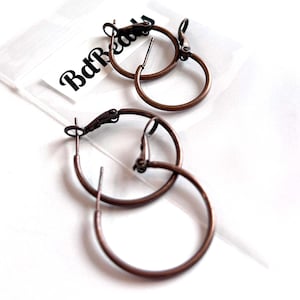 May include: Two pairs of bronze-colored hoop earrings. The earrings are circular with a hinged closure. The image includes a small, clear plastic bag with the text "BdBeads" printed on it. The earrings are displayed on a white surface.
