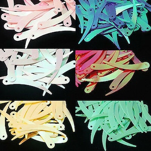 May include: Assortment of colorful, iridescent, curved sequins in various shades of pink, purple, red, yellow, and green. The sequins are arranged in piles, with each pile showcasing a different color.