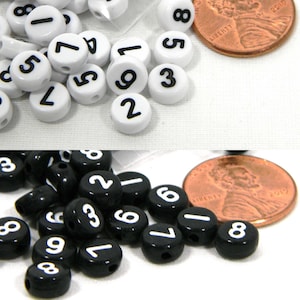 Può includere: Due set di perline rotonde di plastica numerate. Un set è bianco con numeri neri, e l'altro set è nero con numeri bianchi. Le perline sono disposte in un mucchio su una superficie bianca.