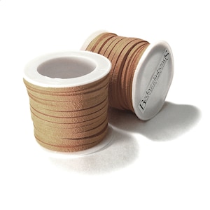 May include: Two spools of tan-colored suede leather cord. The cord is flat and appears to be approximately 3mm wide. The spools have white plastic cores. One spool is upright, and the other is tilted, revealing the brand name.
