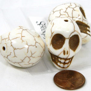 May include: Three white skull beads with brown details. The skulls have a cracked, textured surface. The beads have a hole at the top for stringing.
