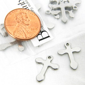 May include: Silver metal cross charms with a hole at the top for hanging. The charms are shown next to a US penny for size comparison.