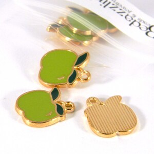 May include: A set of four gold-toned apple charms. Two of the charms are green with a gold outline and green leaves. The other two charms are gold with a textured surface. The charms have a loop at the top for attaching to jewellery or other crafts.