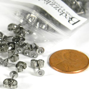 May include: A collection of silver-colored earring posts and backs, scattered on a white surface. A penny is next to the earring components for size comparison. A clear plastic bag with text is in the background.