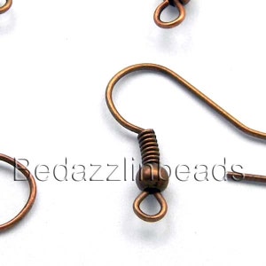 Könnte beinhalten: Nahaufnahme von mehreren kupferfarbenen Ohrringhaken. Jeder Haken hat ein gebogenes Drahtdesign mit einem spiralförmigen Federdetail und einer kleinen Schlaufe am unteren Ende. Der Text "Bedazzlinbeads" ist teilweise sichtbar.