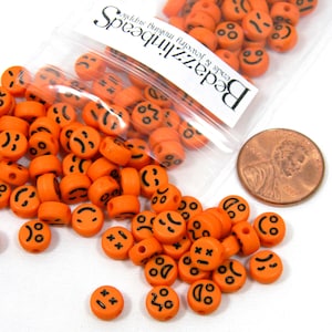 Op de afbeelding: Oranje plastic kralen met verschillende zwarte smiley-gezichtsuitdrukkingen. De kralen zitten in een doorzichtige plastic zak met de tekst "Beadzilli" en "beads & jewelry findings, endless fun!" erop gedrukt. Een cent is opgenomen ter groottevergelijking.