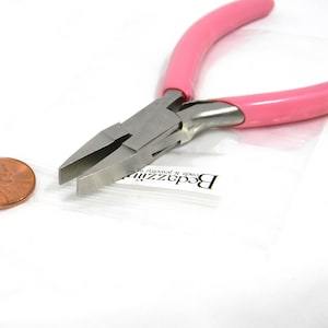 May include: A pair of jewelry pliers with a silver metal head and pink handles. The pliers are open, showing the flat, parallel jaws. A penny is next to the pliers for size comparison. The pliers are on a white surface.