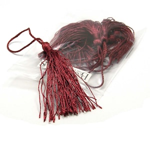 Puede incluir: Un primer plano de un borla de color burdeos con un lazo para colgar. La borla está en una bolsa de plástico transparente con una etiqueta blanca que dice "Beads & More".