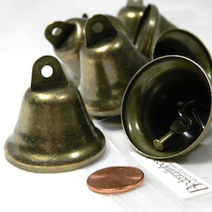 May include: A group of six antique gold-toned jingle bells of varying sizes. The bells are arranged in a semi-circle on a white surface. A penny is in the foreground for scale.