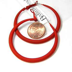 May include: A pair of large red hoop earrings with a clip-on closure. The earrings are shown next to a US penny for size reference.