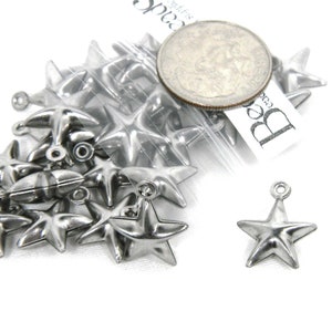 Könnte beinhalten: Eine Tüte mit silbernen, sternförmigen Charms aus Metall, mit einer US-amerikanischen 25-Cent-Münze als Größenvergleich. Die Charms haben oben eine Öse zum Anbringen an Schmuck oder andere Bastelarbeiten.