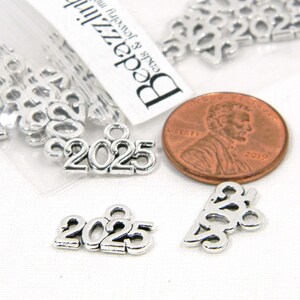 May include: Silver charm with the year 2025, shown next to a US penny for size comparison. The charm is a popular choice for graduation gifts or to commemorate a special year.