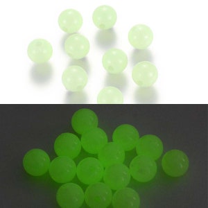 May include: A collection of pale green, round beads with small holes, scattered on a white surface. Below, the same beads glow brightly in a vibrant green against a dark background. These beads are likely for crafting or jewelry making.