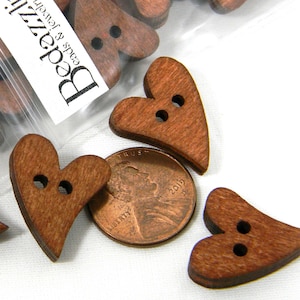 May include: A close-up of a group of brown wooden heart-shaped buttons with two holes each. The buttons are scattered on a white surface next to a penny.