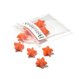 以下が含まれることがあります： オレンジ色の星型のビーズがいくつか入った透明なビニール袋。ビーズは白い背景の上に袋の周りに散らばっています。袋には「Bedazzlin' Beads beads & jewelry making supplies」と書かれたラベルが付いています。