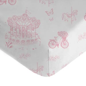 Puede incluir: Sábana de cuna rosa y blanca con un patrón repetido de un carrusel, una bicicleta con flores en la cesta, mariposas y ramitas florales.