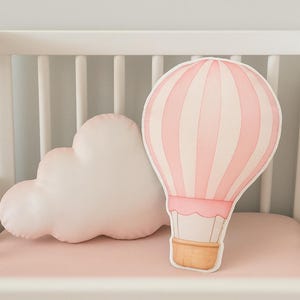 Könnte beinhalten: Ein rosa-weißes Heißluftballon-Kissen und ein wolkenförmiges Kissen liegen auf einer rosa Matratze. Der Heißluftballon hat vertikale Streifen und einen braunen Korb. Das Kinderbett ist weiß.