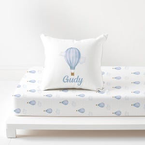 May include: A white pillow and fitted sheet set featuring a blue hot air balloon design. The pillow has a watercolor balloon with the name "Gudy" printed below. The fitted sheet has a repeating pattern of blue balloons and clouds.