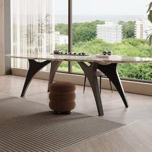 Op de afbeelding: Een ovale marmeren eettafel met donkerbruine poten. De tafel is gedekt met een theeset en decoratieve items. De kamer heeft een groot raam met uitzicht op bomen en gebouwen. Een bruin tapijt en een bruine poef staan op de voorgrond.