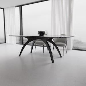 May include: A modern black dining table with a marble top and unique, curved legs. The table is in a bright, minimalist room with two gray chairs and a decorative black bowl. The room has large windows with white curtains.