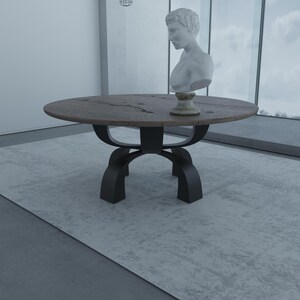 May include: A round wooden dining table with a dark brown top and a black metal base. The table has a unique, curved base design. A white plaster bust of a man is sitting on the table.