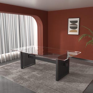 Op de afbeelding: Een moderne eettafel met een helder glazen blad en een donkergrijze metalen voet. De voet heeft verticale lamellen. De tafel staat op een grijs patroonkleed. De kamer heeft een roodbruine muur en een raam met witte gordijnen.