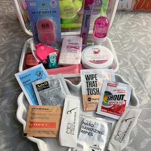 May include: A clear iridescent travel case filled with various personal care items including AOA soap sheets, a Bloom watermelon stick, a pink mirror, Marie Claire pocket tissues, Dove cream, Advil, Biopure wipes, Shout wipes, Brookstone makeup remover, and dental floss picks.