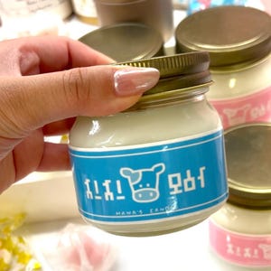 Puede incluir: Una peque&ntilde;a vela en tarro de cristal con tapa dorada y etiqueta azul con un gr&aacute;fico de vaca y texto. La vela tiene cera blanca y la sostiene una mano. Se ven otras velas en el fondo.
