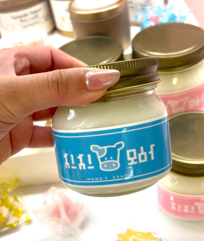 Puede incluir: Una peque&ntilde;a vela en tarro de cristal con tapa dorada y etiqueta azul con un gr&aacute;fico de vaca y texto. La vela tiene cera blanca y la sostiene una mano. Se ven otras velas en el fondo.