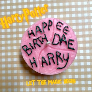 Pode incluir: Vela de aniversário rosa com as palavras "Happy Birthday Harry" escritas em preto. A vela está sobre um fundo xadrez com as palavras "Let the Magic Begin" escritas em amarelo.