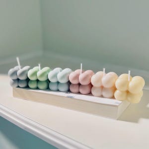 Könnte beinhalten: Eine Reihe von pastellfarbenen Blasenkerzen, die auf einer weißen Oberfläche angeordnet sind. Die Kerzen sind in den Farben Grün, Blau, Rosa und Gelb.