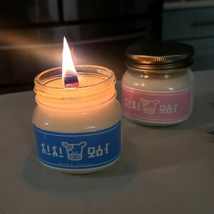 Puede incluir: Dos velas encendidas en peque&ntilde;os frascos de vidrio. La vela encendida tiene una etiqueta azul con texto coreano y un gr&aacute;fico de vaca. La otra vela tiene una etiqueta rosa con texto coreano y un gr&aacute;fico de vaca.