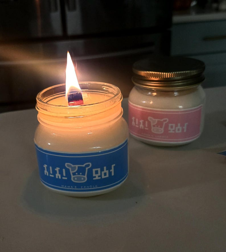 Puede incluir: Dos velas encendidas en peque&ntilde;os frascos de vidrio. La vela encendida tiene una etiqueta azul con texto coreano y un gr&aacute;fico de vaca. La otra vela tiene una etiqueta rosa con texto coreano y un gr&aacute;fico de vaca.