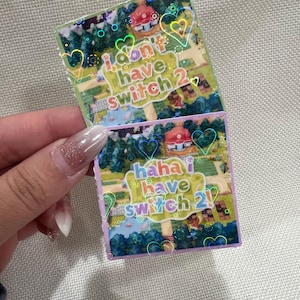 Puede incluir: Dos pegatinas holográficas con un diseño de paisaje colorido y texto. Una pegatina dice "I don't have Switch 2" y la otra dice "haha I have Switch 2!" con acentos de corazón y círculo.