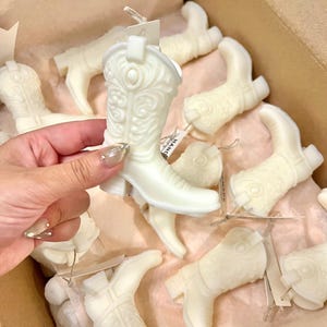 May include: Ivory-colored cowboy boot-shaped candles with intricate embossed designs. The boots are arranged in a cardboard box, some with tags attached. The candles have a Western theme.