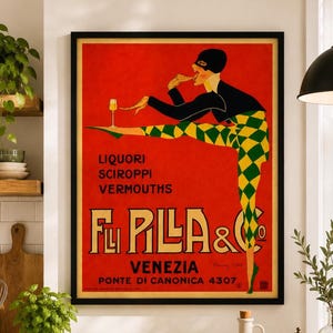 Può includere: Un poster incorniciato in stile vintage su sfondo rosso raffigurante una figura con maschera nera e abito a rombi neri e gialli. La figura bilancia un bicchiere su un piede. Testo: LIQUORI SCIROPPI VERMOUTHS FLLI PILLA & CO VENEZIA.