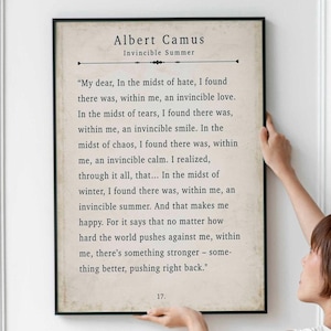 May include: A framed print featuring a quote by Albert Camus, titled "Invincible Summer." The text is in a classic serif font on a cream-colored background. The frame is black, and the print is held by a person.