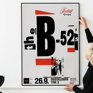 Könnte beinhalten: Schwarz-weißes Poster mit roten Akzenten, das ein Konzert von The B-52's bewirbt. Das Poster zeigt den Bandnamen in großen, fetten Buchstaben und ein kleineres Bild der Bandmitglieder. Der Text auf dem Poster lautet "The B-52's" und "Dusseldorf Tor 3".