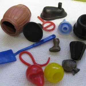 Può includere: Una collezione di giocattoli di plastica vintage di vari colori, tra cui una botte marrone, uno stivale nero, una pala blu, un gancio rosso, una campana gialla e rossa, un cappello nero e un pesce nero e grigio.