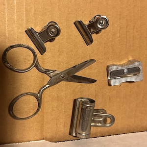 May include: An assortment of silver-colored office supplies, including three binder clips, a pair of small scissors, and a pencil sharpener. The items are arranged on a brown cardboard surface.