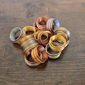 May include: A collection of 15 decorative rings in gold, silver, and copper tones. The rings are stacked and arranged in a pile. Each ring has a different pattern of stripes.