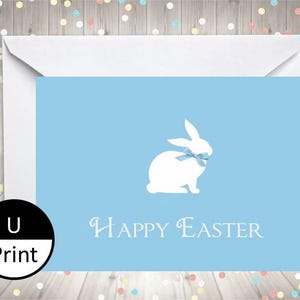 Könnte beinhalten: Hellblaue Ostergrußkarte mit einer weißen Hasenillustration und dem Schriftzug "Happy Easter". Die Karte befindet sich in einem weißen Umschlag. Der Hintergrund ist Holz mit Konfetti.