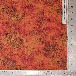 May include: A piece of fabric with an abstract pattern in shades of orange, red, and brown. The fabric has a mottled appearance, with darker spots scattered throughout. The text "Autumn Glory by Freckle + Lollie" is visible on a white label.